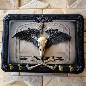 May include: A black framed key holder with a skull and crossbones design. The skull is surrounded by black wings and a gold heart with a skull and crossbones design. The key holder has five hooks.