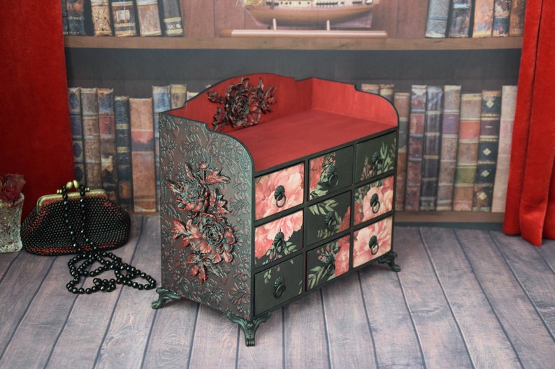 May include: A black wooden drawer organizer with a red top and floral patterned drawers. The organizer has a floral design on the front and is decorated with a 3D floral accent.