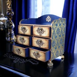 May include: A small wooden chest with six drawers, each with a decorative gold knob. The chest is painted blue and gold and has a damask pattern on the side.