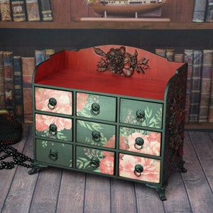 May include: A small black wooden chest with intricate carvings and floral details. The chest has nine drawers with floral patterns and black metal handles. The top of the chest is red and has a carved floral design.
