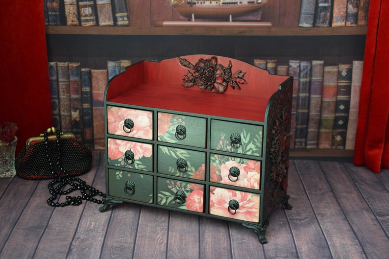May include: A small black wooden chest with intricate carvings and floral details. The chest has nine drawers with floral patterns and black metal handles. The top of the chest is red and has a carved floral design.