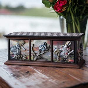 Op de afbeelding: Een houten doos met drie laden met illustraties met het thema Alice in Wonderland. De laden zijn versierd met afbeeldingen van Alice, de Cheshire Cat en het Witte Konijn.