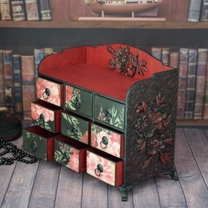 May include: A black wooden decorative box with eight drawers. The drawers are decorated with floral patterns in shades of pink and green. The box has a red velvet interior and intricate floral carvings on the sides.