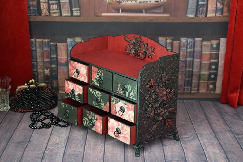 May include: A black wooden decorative box with eight drawers. The drawers are decorated with floral patterns in shades of pink and green. The box has a red velvet interior and intricate floral carvings on the sides.