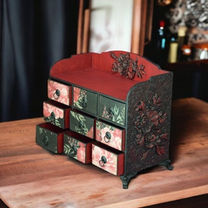 May include: A small wooden chest with eight drawers, each with a floral design. The chest is painted black and red and has ornate floral carvings on the sides. The chest is sitting on a wooden table.