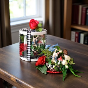 Puede incluir: Una caja de regalo cilíndrica transparente con una cinta de cuadros blancos y negros que contiene una rosa roja y una figura de conejo, junto a un arreglo abierto con una figura de conejo, rosas rojas y blancas, flores azules y una taza a cuadros.
