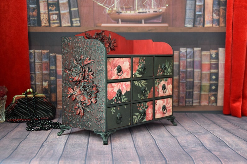 May include: A black wooden drawer organizer with a red top and floral patterned drawers. The organizer has decorative floral carvings and sits on four legs.