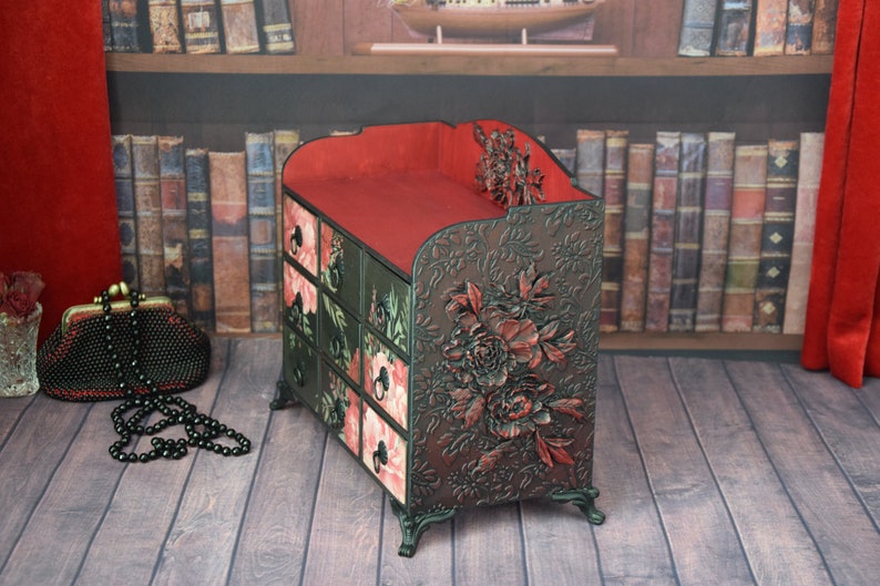 May include: A small, multi-drawered wooden box with a red top and floral carvings. The box is decorated with a floral pattern and has a dark brown finish.