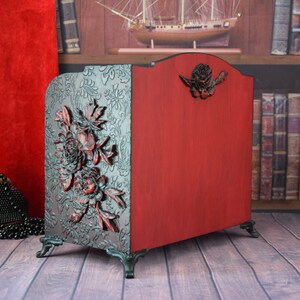 May include: A red wooden magazine rack with carved floral details and black metal feet. The rack is decorated with a floral design and has a red finish.
