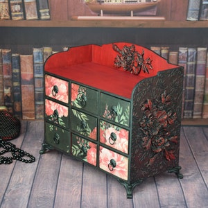 May include: A black wooden decorative box with eight drawers. The drawers are decorated with floral patterns in shades of pink, green, and black. The top of the box is red and has a floral design. The box is sitting on a wooden floor.