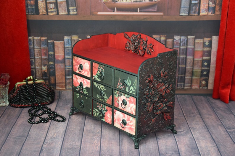 May include: A black wooden decorative box with eight drawers. The drawers are decorated with floral patterns in shades of pink, green, and black. The top of the box is red and has a floral design. The box is sitting on a wooden floor.