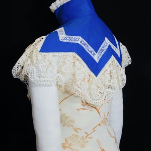 May include: A white lace collar with a blue fabric insert. The collar is trimmed with white lace and has a scalloped edge. The fabric insert is a bright blue colour and has a white lace trim.