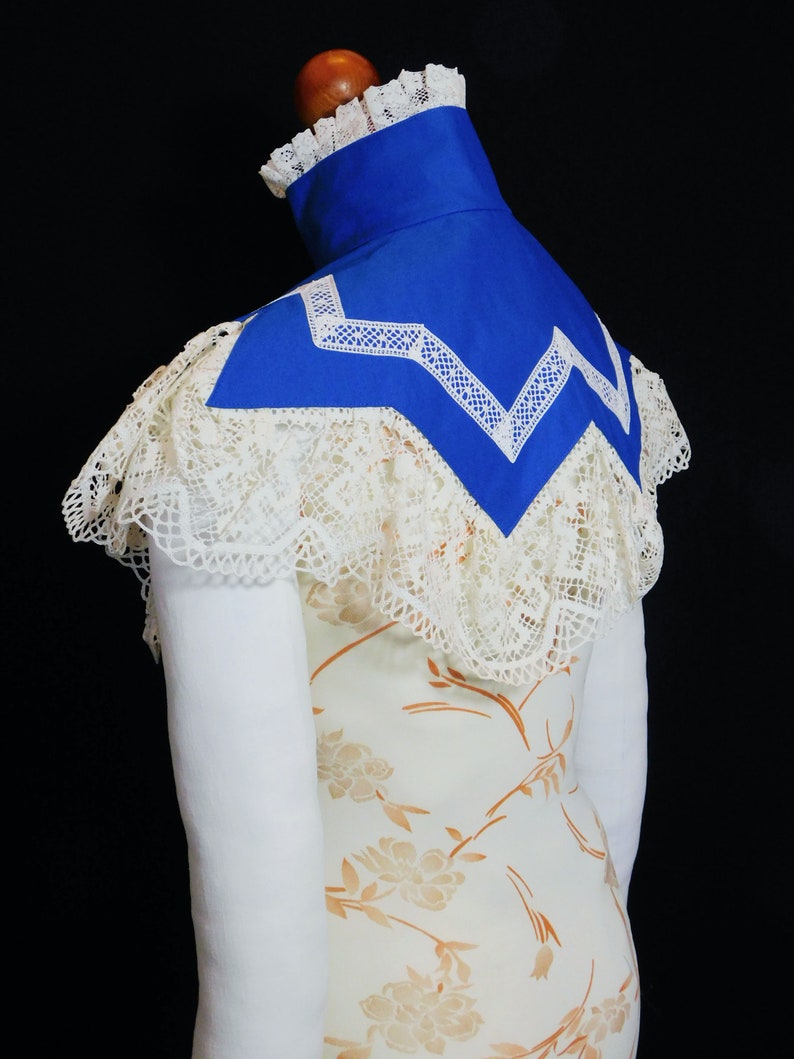 May include: A white lace collar with a blue fabric insert. The collar is trimmed with white lace and has a scalloped edge. The fabric insert is a bright blue colour and has a white lace trim.