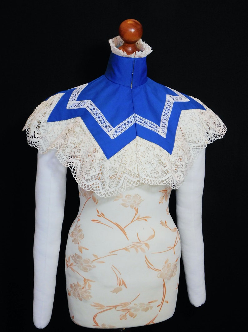May include: A white lace collar with a blue fabric insert. The collar is on a white dress form with long white sleeves.