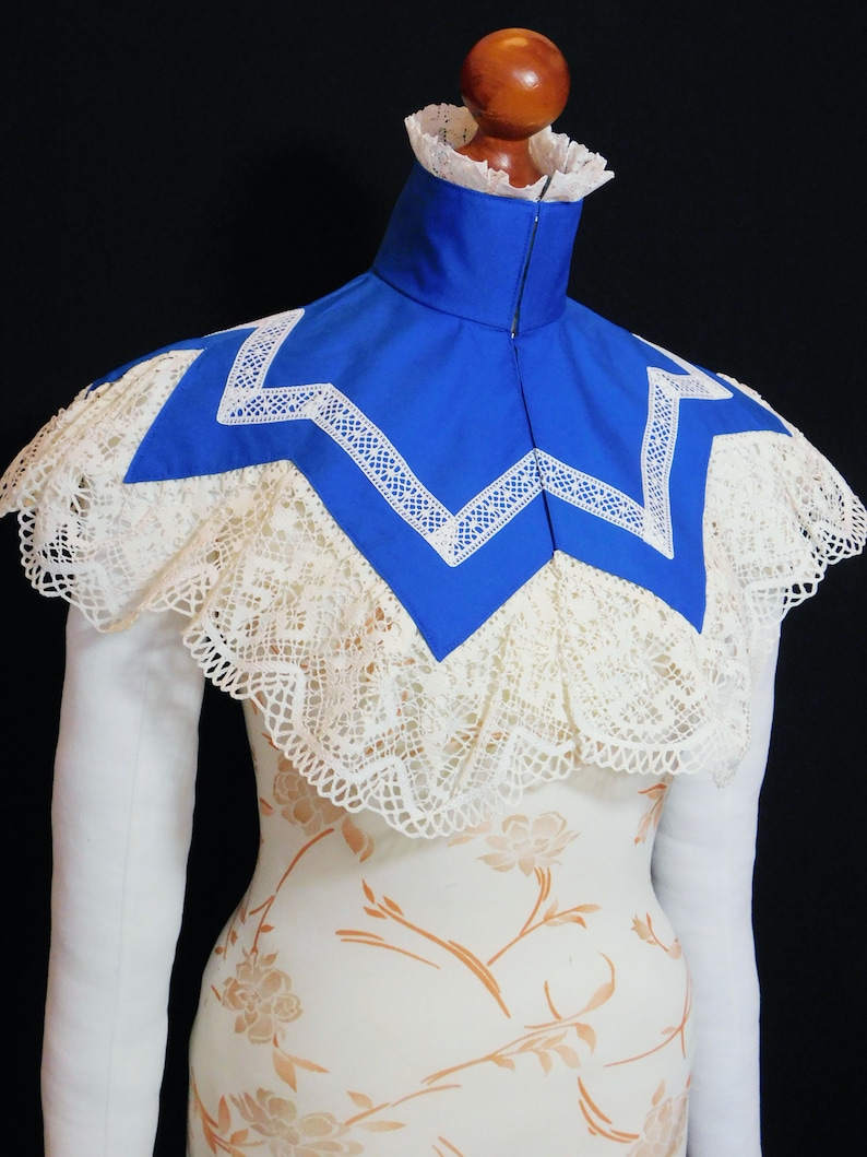 May include: A blue lace collar with a white lace trim. The collar is on a white dress form with a floral pattern.