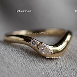 May include: A gold-colored V-shaped ring with three small, clear gemstones. The ring has a smooth, polished finish and is set against a soft, neutral background. The brand name "Fantique Jewels" is visible in the image.