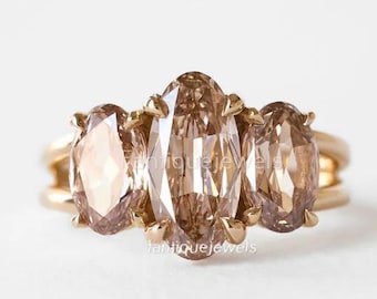 May include: A gold ring featuring three oval-cut champagne-colored gemstones set in a horizontal row. The center stone is larger than the two flanking stones, and all are secured by gold prongs on a simple, polished gold band. The logo Fantique Jewels is visible.