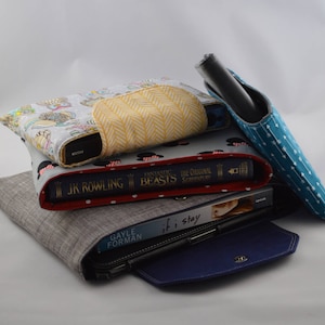 May include: A stack of books and a blue fabric case with a zipper. The top book is a red hardback with the title "Fantastic Beasts The Original Screenplay" by J.K. Rowling. The second book is a black hardback with the title "If I Stay" by Gayle Forman. The third book is a grey hardback with a blue fabric case on top.