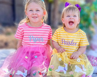 May include: Two girls wearing pink and yellow striped dresses with tulle skirts and embroidered bunnies and flowers. The girl on the left's dress says "Piper" and the girl on the right's dress says "Paislee".