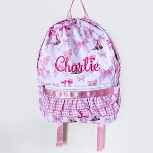 May include: A white backpack featuring a repeating pattern of pink bows and small brown boots. The name Charlie is embroidered in pink script across the center. The bag includes a front zippered pocket with a pink ribbon and a ruffled gingham trim.