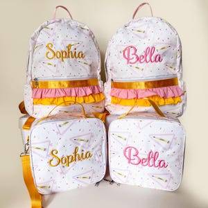 Pode incluir: Mochilas e lancheiras brancas com um padrão de lápis e triângulos. As mochilas têm acabamentos dourados, babados rosa e amarelos e os nomes "Sophia" e "Bella" bordados em ouro e rosa. As lancheiras têm nomes correspondentes.