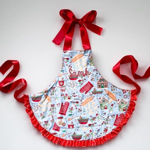 May include: A blue and red Christmas-themed apron with a pocket and a red ribbon tie. The apron has a gingerbread house, snowman, and other holiday-themed designs. The name "Sarah" is embroidered on the apron.