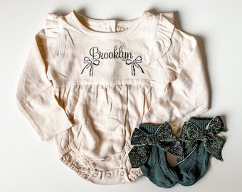May include: A cream-colored baby onesie with long sleeves and a ruffled neckline. The onesie has the name "Brooklyn" embroidered on the front with two bows on either side of the name. The onesie is paired with a pair of dark green socks with a sparkly bow on each sock.
