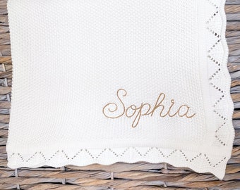May include: White knitted baby blanket with a delicate lace border and the name "Sophia" embroidered in gold thread.