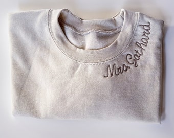 May include: A light beige sweatshirt with the text "Mrs. Got Hand" embroidered on the neckline.