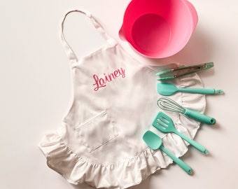 May include: A white apron with a ruffled hem and a pocket. The apron is embroidered with the name "Lainey" in pink. The apron is accompanied by a pink mixing bowl and a set of turquoise kitchen utensils.