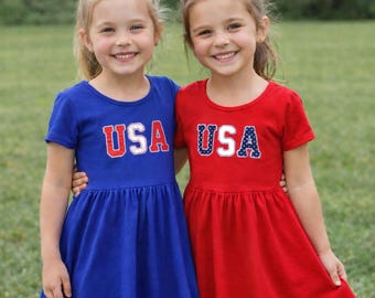 May include: Two young girls wearing matching dresses. One dress is royal blue, the other is red. Both dresses have the word "USA" in a patriotic design on the front. The girls are smiling and standing on a grassy field.