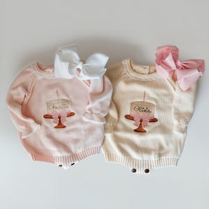 Könnte beinhalten: Zwei Baby-Strampler, einer hellrosa und einer cremefarben, jeweils mit einem auf der Vorderseite gestickten Kuchenmotiv. Der Kuchen hat eine rosa Schleife und eine Kerze. Der rosa Strampler hat eine weiße Schleife, und der cremefarbene Strampler hat eine rosa Schleife. Der rosa Strampler sagt "Charlie" und der cremefarbene Strampler sagt "Olivia".