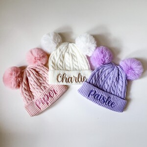 Könnte beinhalten: Drei gestrickte Baby-Mützen mit Bommeln. Die Mützen sind weiß, rosa und lila. Jede Mütze hat einen Namen auf der Vorderseite gestickt: Charlie, Piper und Paislee.