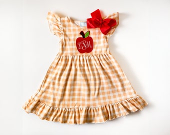 Puede incluir: Un vestido de cuadros vichy amarillo y blanco con una manzana roja bordada en la parte delantera. La manzana tiene las letras "LSM" bordadas. El vestido tiene un dobladillo con volantes y mangas cortas. Un lazo rojo está en la parte superior del vestido.