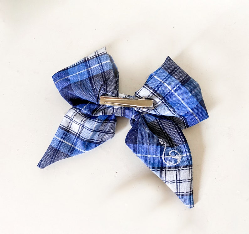 May include: A blue and white plaid bow with a silver clip. The bow has a small embroidered letter "S" on one side.