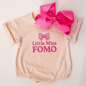 May include: A light peach-colored baby romper with rolled sleeves and the text "Little Miss FOMO" in pink. A large, bright pink bow is attached to the top of the romper.