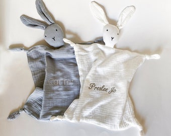 May include: Two personalized baby security blankets with bunny head attachments. The blanket on the left is gray with the name "Carter" embroidered on it. The blanket on the right is white with the name "Preslee Jo" embroidered on it.