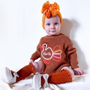 Pode incluir: Um bebê vestindo um body de malha marrom com um desenho de peru e o nome "Charlie" bordado. O bebê também está usando uma faixa de cabelo com laço laranja, meias laranja e sapatos brancos com laços.