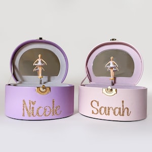 Puede incluir: Dos cajas de música ovaladas abiertas, una lavanda y otra rosa, cada una con un espejo y una figura de bailarina. La caja lavanda tiene el nombre "Nicole" en purpurina dorada, y la caja rosa tiene "Sarah". Ambas tienen cierres dorados.