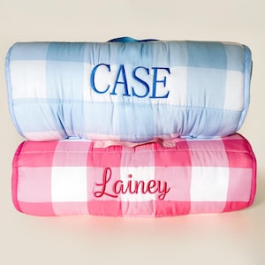 May include: Two rolled-up, padded blankets. The top blanket is light blue and white checkered, embroidered with the name "CASE" in blue. The bottom blanket is pink and white checkered, embroidered with the name "Lainey" in pink.