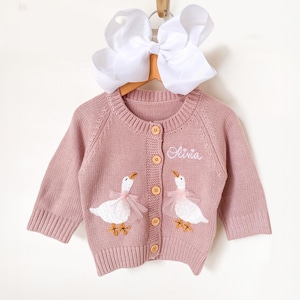 Könnte beinhalten: Ein staubrosa Baby-Cardigan mit rundem Ausschnitt und langen Ärmeln. Der Pullover hat zwei gestickte weiße Gänse mit rosa Tüllakzenten und Holzknöpfen. Der Name "Olivia" ist auf der Vorderseite gestickt.