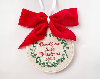 May include: A round embroidery hoop ornament featuring a cream-colored fabric center with green and red embroidered holly wreath detailing. The text reads Brooklyn's first Christmas 2026. A large red velvet bow is attached to the top with a white ribbon.