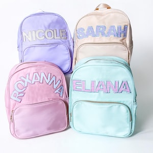 Op de afbeelding: Vier kleurrijke rugzakken met gepersonaliseerde namen in een ruitjespatroon. De rugzakken zijn paars, beige, roze en lichtblauw.