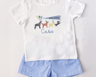 May include: A white t-shirt and blue gingham shorts set. The shirt features an embroidered design of dogs with balloons and a banner that says "Happy Birthday". The name "Case" is embroidered below the dogs.