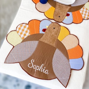May include: A fabric turkey-shaped placemat featuring a brown corduroy head and body with the name Sophia embroidered in white script. The tail feathers consist of various patterned fabric segments in orange, yellow, blue, and white gingham checks.