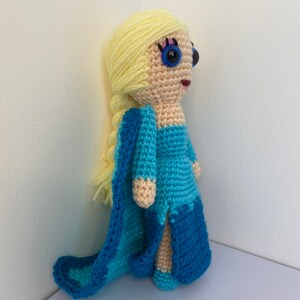 K&ouml;nnte beinhalten: Eine geh&auml;kelte Puppe mit langen blonden Haaren, die ein blaues Kleid und einen passenden Umhang tr&auml;gt. Die Puppe hat blaue Augen und einen rosa Mund.