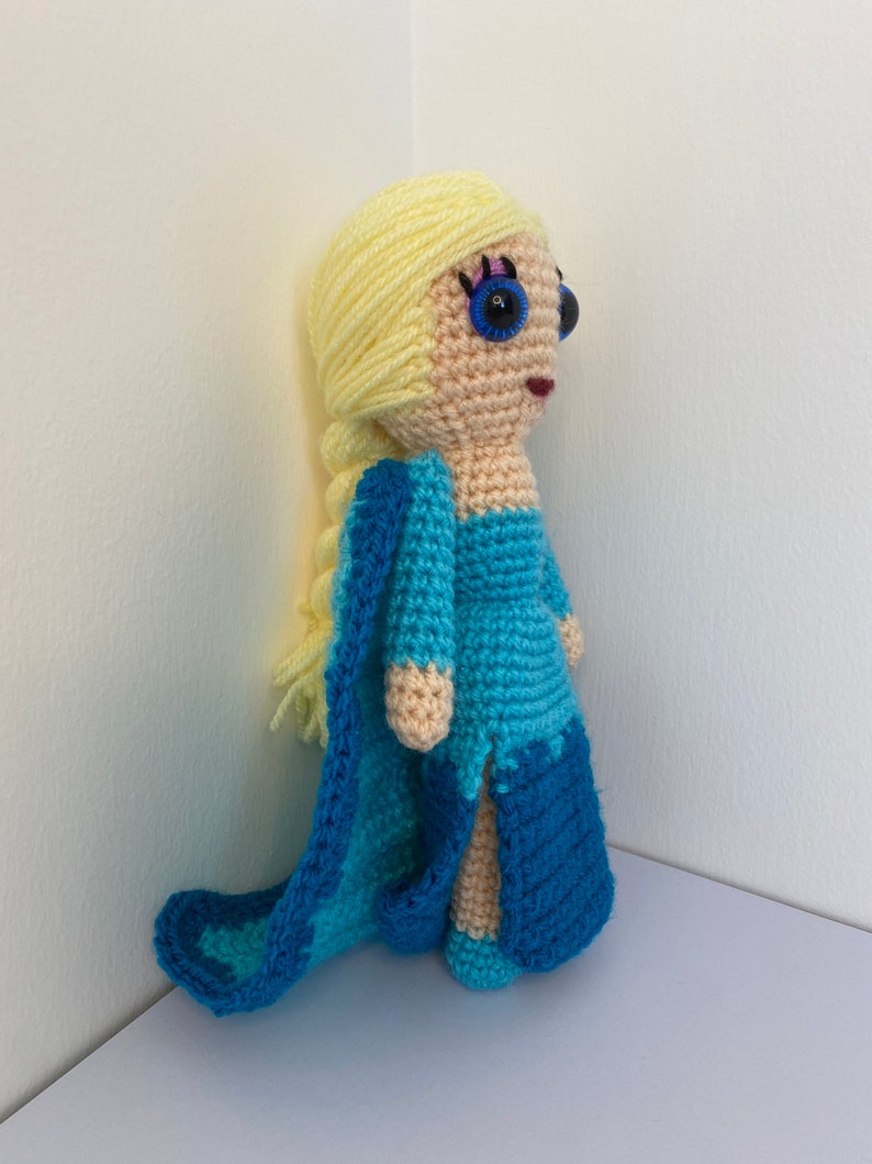 K&ouml;nnte beinhalten: Eine geh&auml;kelte Puppe mit langen blonden Haaren, die ein blaues Kleid und einen passenden Umhang tr&auml;gt. Die Puppe hat blaue Augen und einen rosa Mund.