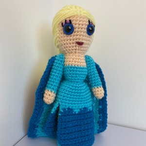 K&ouml;nnte beinhalten: Eine geh&auml;kelte Puppe mit blonden Haaren, blauen Augen und einem blauen Kleid. Die Puppe tr&auml;gt einen blauen Umhang.