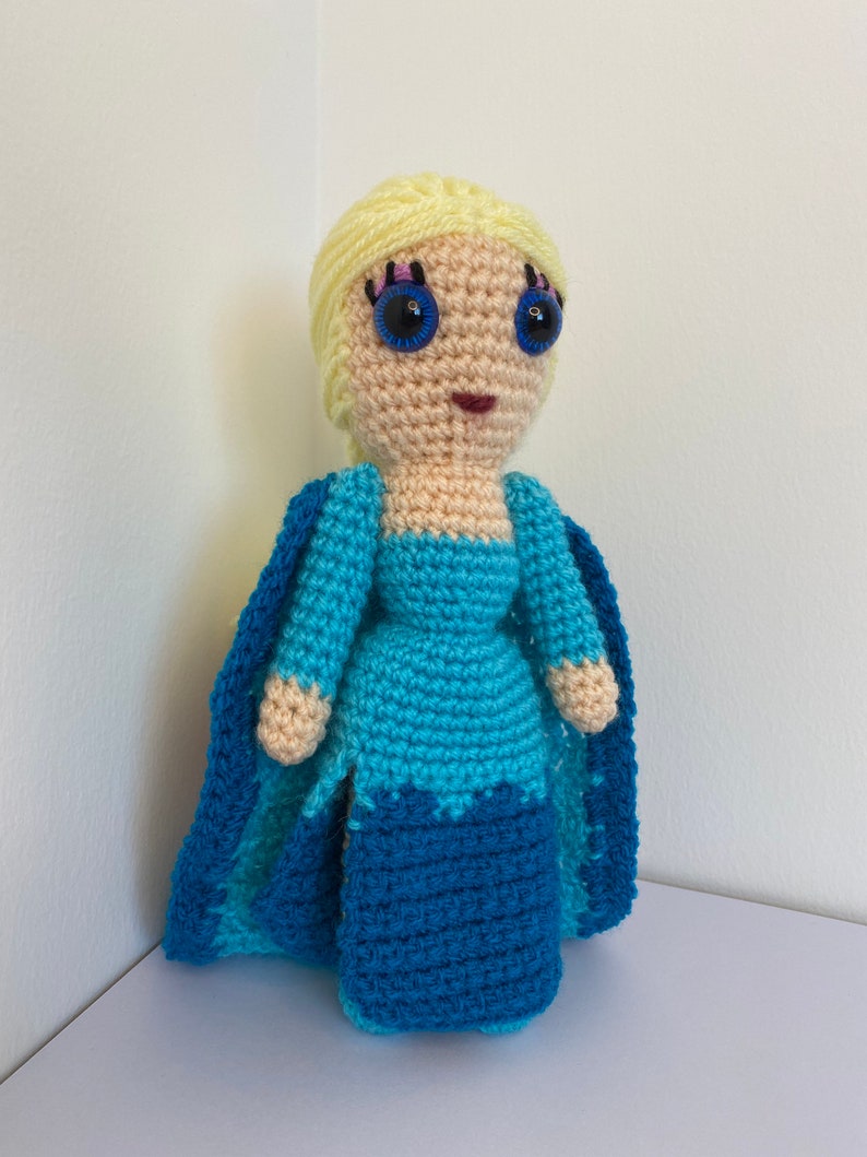 K&ouml;nnte beinhalten: Eine geh&auml;kelte Puppe mit blonden Haaren, blauen Augen und einem blauen Kleid. Die Puppe tr&auml;gt einen blauen Umhang.