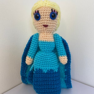 K&ouml;nnte beinhalten: Eine geh&auml;kelte Puppe mit blonden Haaren, blauen Augen und einem blauen Kleid. Die Puppe tr&auml;gt einen blauen Umhang.